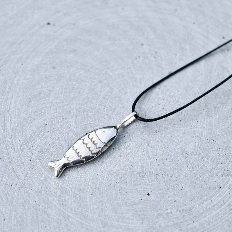 A silver fish-shaped pendant necklace with a black cord, displayed on a textured gray surface.