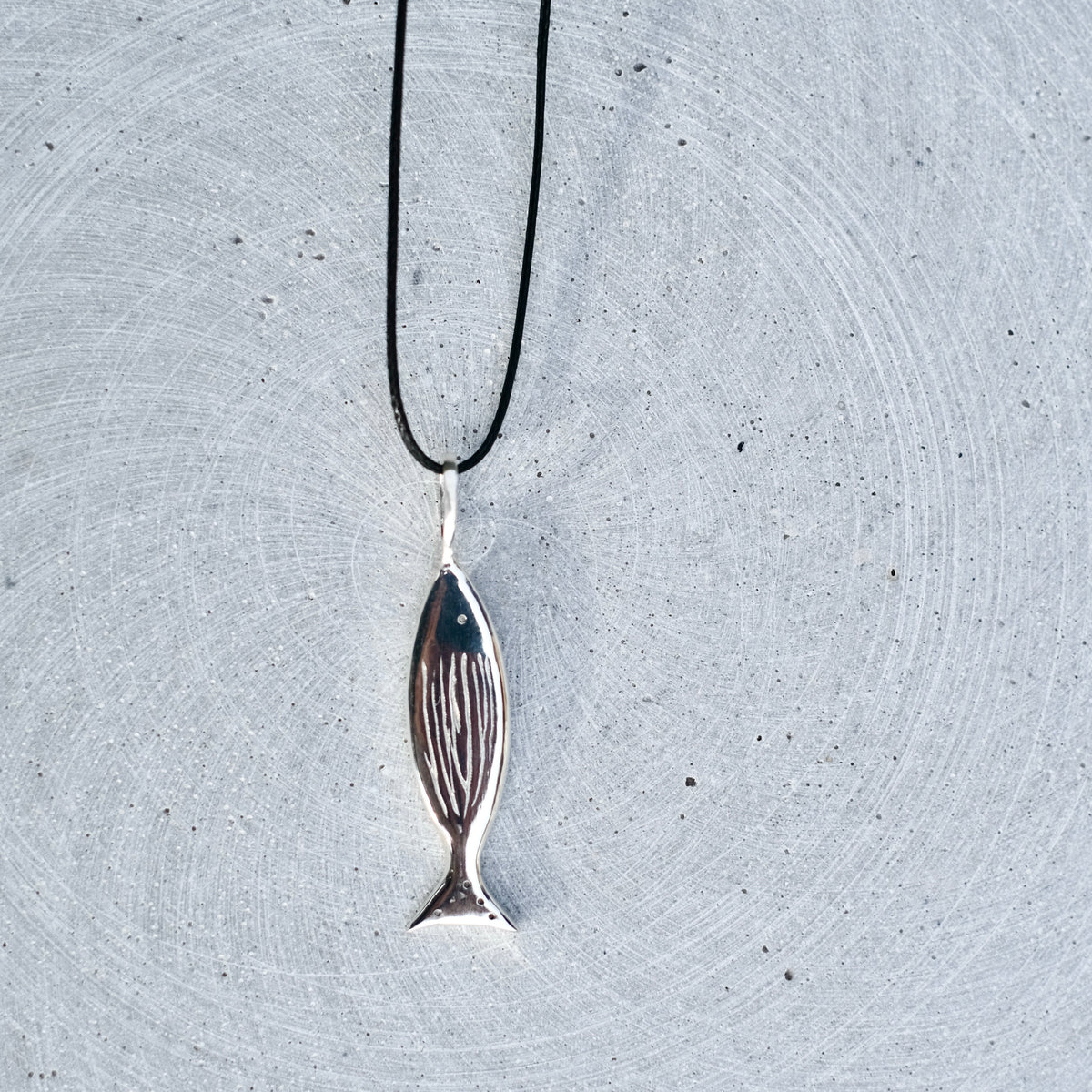 A silver fish pendant necklace with a black cord, laid horizontally on a textured light gray concrete surface.