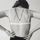 A black and white photo of a woman from behind wearing a minimalist square pendant necklace with a black cord and white square pendant, styled with a white ruffled bra and black shorts.