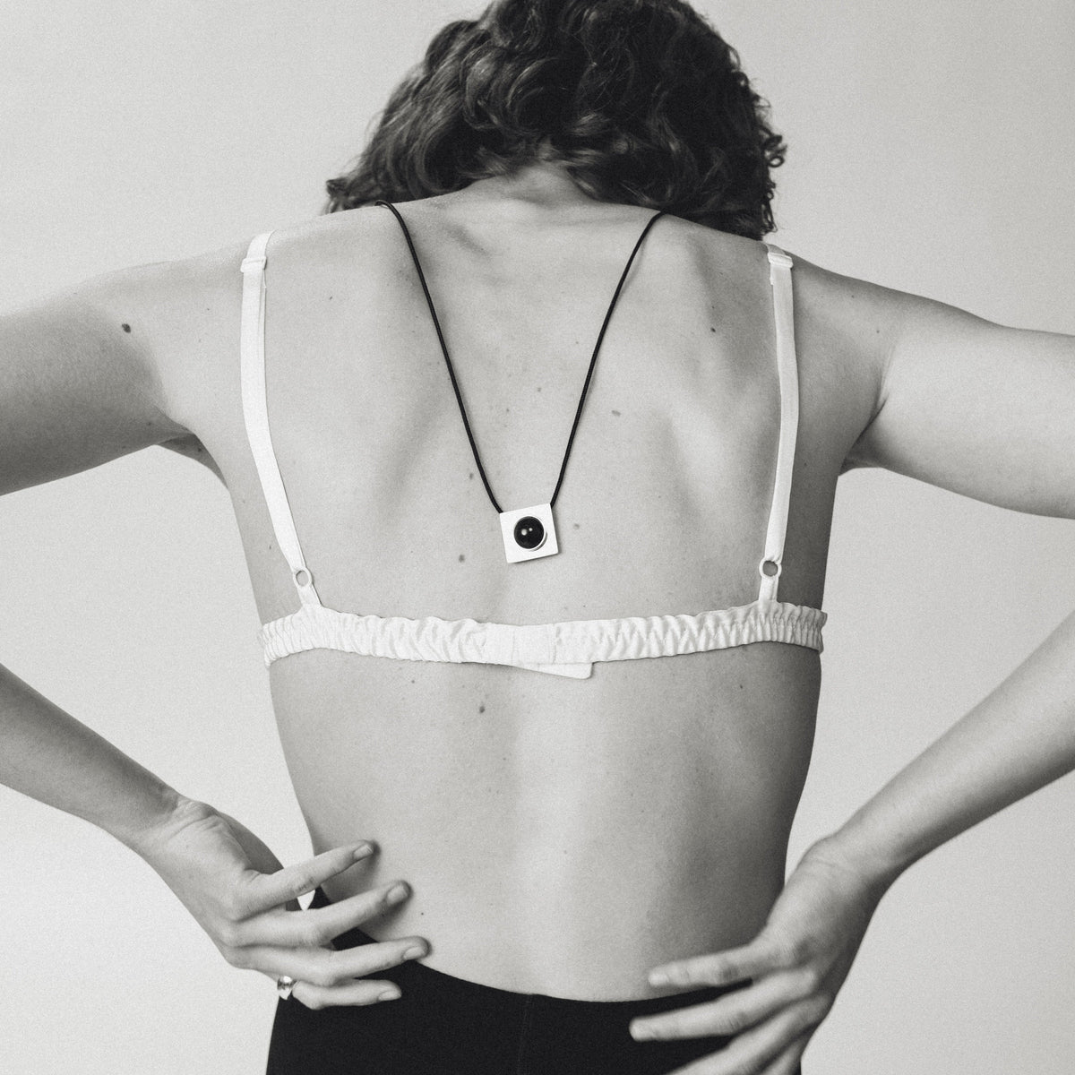 A black and white photo of a woman from behind wearing a minimalist square pendant necklace with a black cord and white square pendant, styled with a white ruffled bra and black shorts.