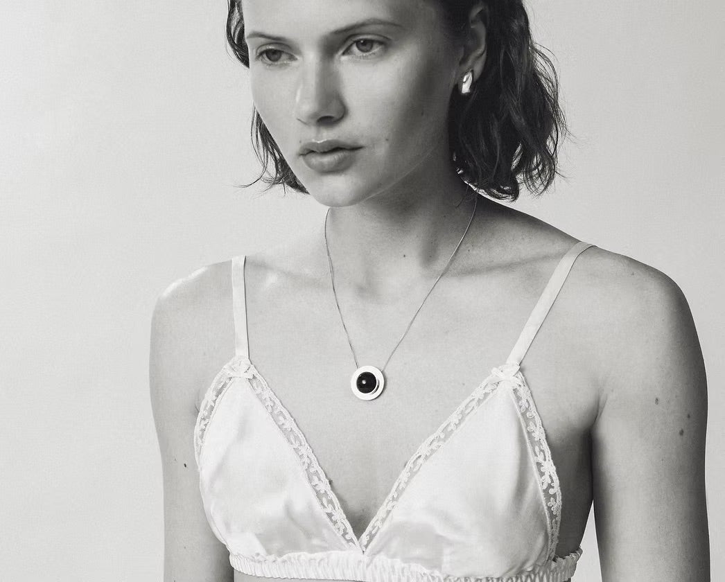 A close-up black and white lifestyle shot of a woman wearing a delicate silver chain necklace with a circular pendant featuring a dark amber at the center.