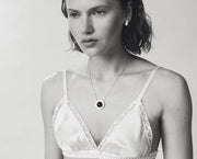 A close-up black and white lifestyle shot of a woman wearing a delicate silver chain necklace with a circular pendant featuring a dark amber at the center.