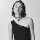 A black one-shoulder button-front vest worn by a model with her eyes closed, paired with a white skirt against a light gray studio background.