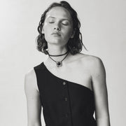 A black one-shoulder button-front vest worn by a model with her eyes closed, paired with a white skirt against a light gray studio background.