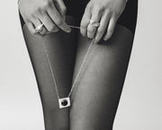 A close-up black and white photo of a person wearing sheer black tights, holding a delicate silver chain necklace with a square pendant featuring a dark circular center. The person is also wearing silver rings.