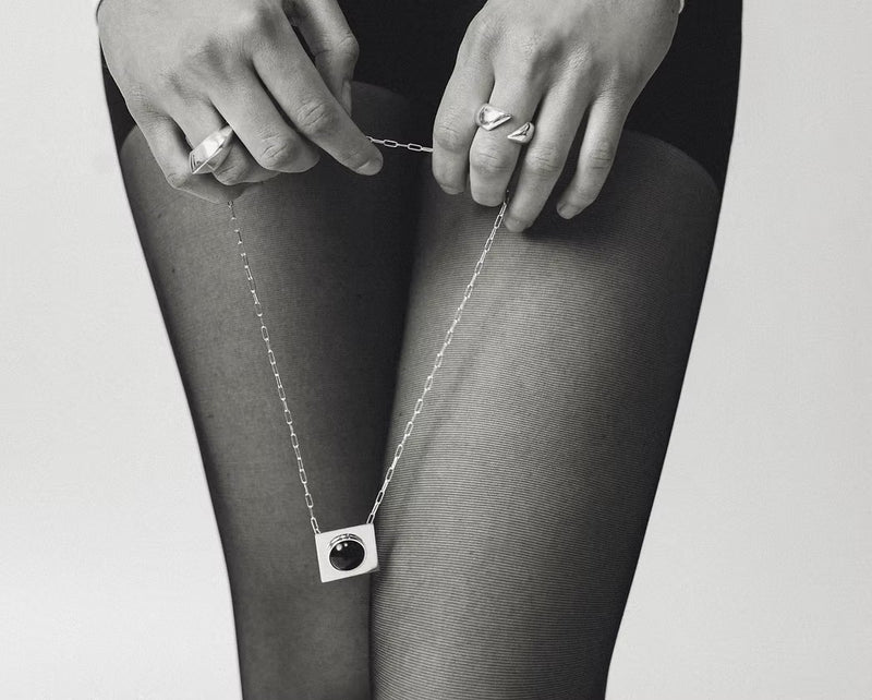 A close-up black and white photo of a person wearing sheer black tights, holding a delicate silver chain necklace with a square pendant featuring a dark circular center. The person is also wearing silver rings.