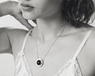 A close-up black and white lifestyle shot of a woman wearing a delicate silver chain necklace with a circular pendant featuring a dark amber at the center.