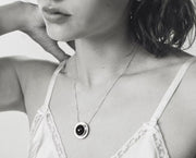 A close-up black and white lifestyle shot of a woman wearing a delicate silver chain necklace with a circular pendant featuring a dark amber at the center.