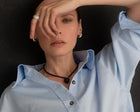 A close-up portrait of a woman wearing a light blue button-up shirt, a black cord necklace with a round black pendant, silver hoop earrings, and silver rings, posed against a dark background.