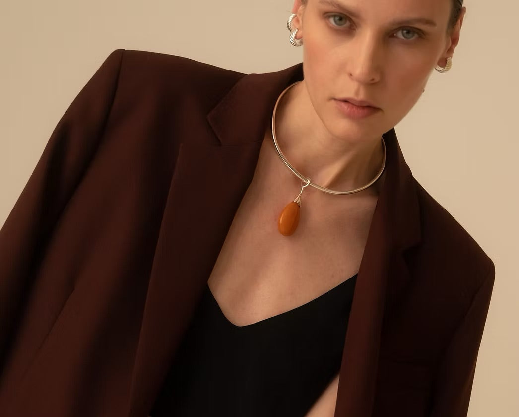 A close-up of a model wearing a silver choker necklace with a smooth, teardrop-shaped amber pendant, styled with a dark brown blazer and black top against a solid beige background.