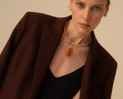 A close-up of a model wearing a silver choker necklace with a smooth, teardrop-shaped amber pendant, styled with a dark brown blazer and black top against a solid beige background.