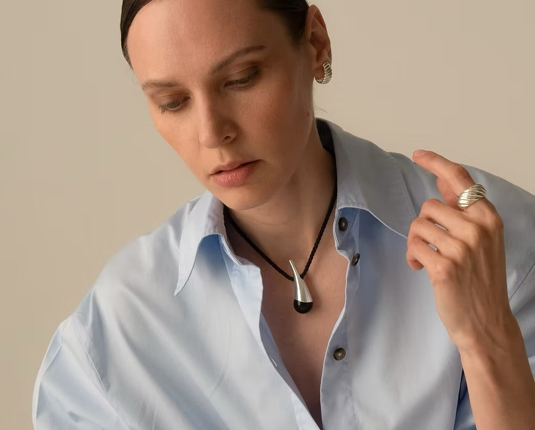 A close-up lifestyle shot of a woman wearing a black cord necklace with a polished silver-toned pendant shaped like a stylized animal skull or horn. She is wearing a light blue button-down shirt and a silver ring on one finger, set against a solid warm beige background.