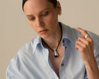A close-up lifestyle shot of a woman wearing a black cord necklace with a polished silver-toned pendant shaped like a stylized animal skull or horn. She is wearing a light blue button-down shirt and a silver ring on one finger, set against a solid warm beige background.