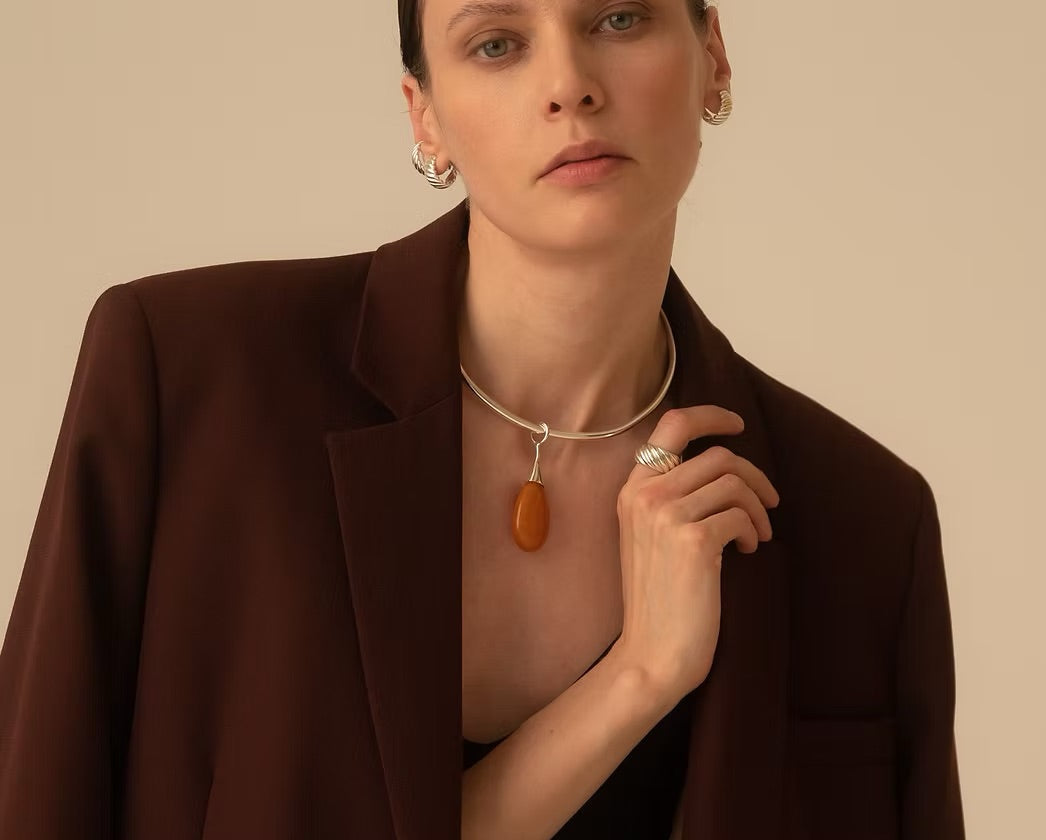 A close-up of a model wearing a silver-toned choker necklace with a teardrop-shaped amber pendant, styled with a dark brown blazer against a solid beige background.