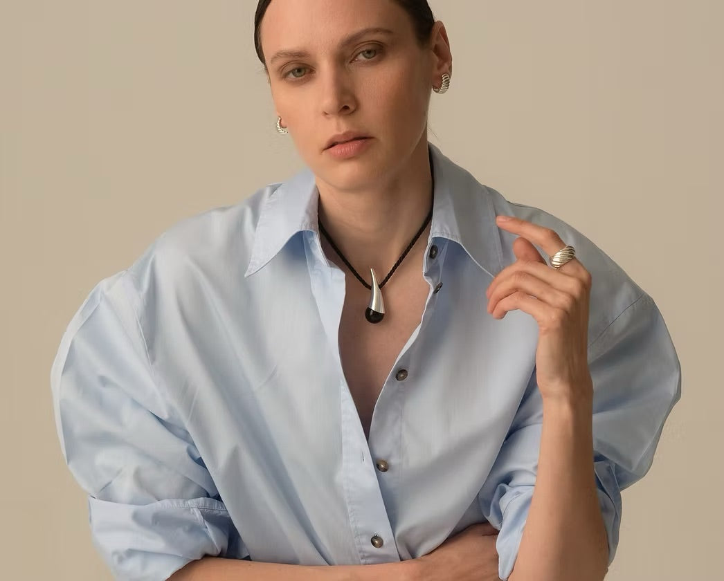A model wearing a light blue oversized button-down shirt and a black cord necklace with a silver-toned teardrop pendant, posed against a solid beige background.