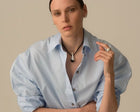 A model wearing a light blue oversized button-down shirt and a black cord necklace with a silver-toned teardrop pendant, posed against a solid beige background.
