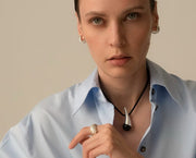 A close-up portrait of a model wearing a silver teardrop pendant necklace on a black cord, a silver ring, and silver hoop earrings, wearing a light blue button-down shirt against a warm beige background.