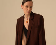 A model wearing a dark brown blazer over a black top, featuring a silver necklace with a teardrop-shaped amber pendant, set against a solid beige background.