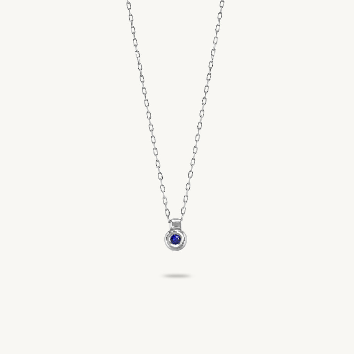 A delicate silver chain necklace with a small circular pendant featuring a deep blue gemstone, displayed against a solid off-white background.