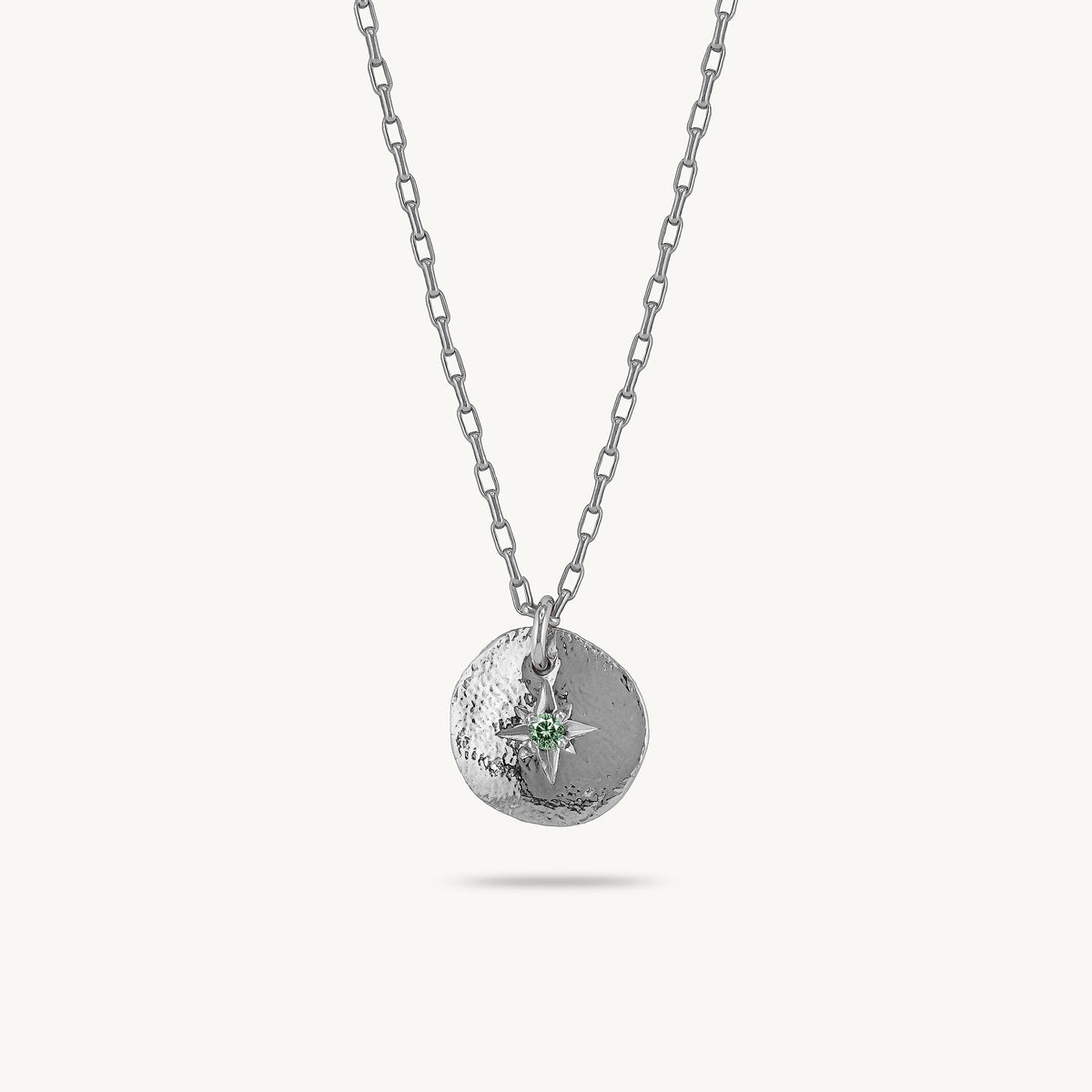 A silver chain necklace with a textured circular pendant featuring a small silver compass rose and a tiny green gemstone, shown against a light off-white background.