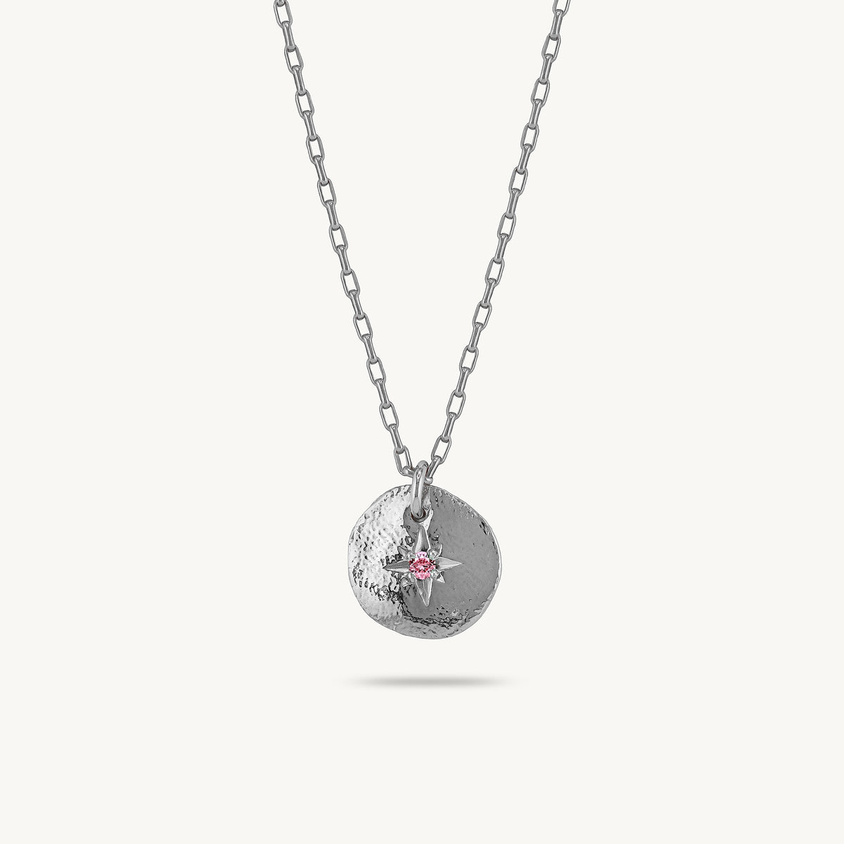 A silver chain necklace with a textured circular pendant featuring a small silver compass rose and a tiny pink sapphire accent, shown against a light off-white background.