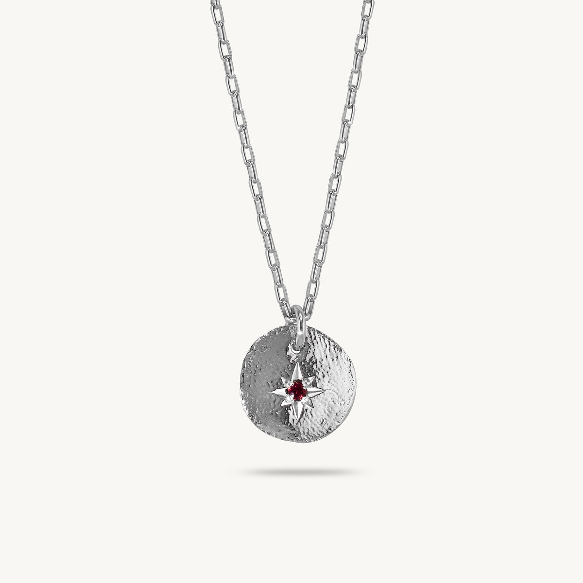 A silver chain necklace with a textured circular pendant featuring a small red sapphire compass star in the center, shown against a light cream background.