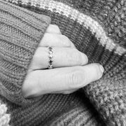 A close-up of a hand wearing a delicate silver ring with a scalloped, wavy edge, resting against a cozy, textured gray and white striped knit sweater.