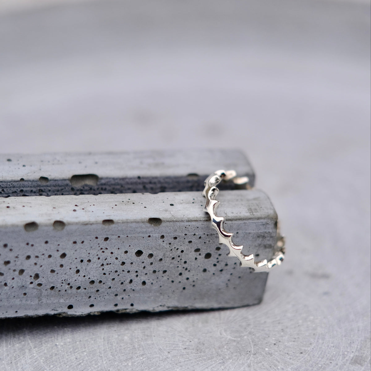 A delicate silver ring with a scalloped, wavy edge, resting on textured gray concrete blocks against a soft gray background.