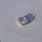 A clear faceted gemstone ring with a textured silver bezel, resting on a light gray textured surface with a sharp shadow.