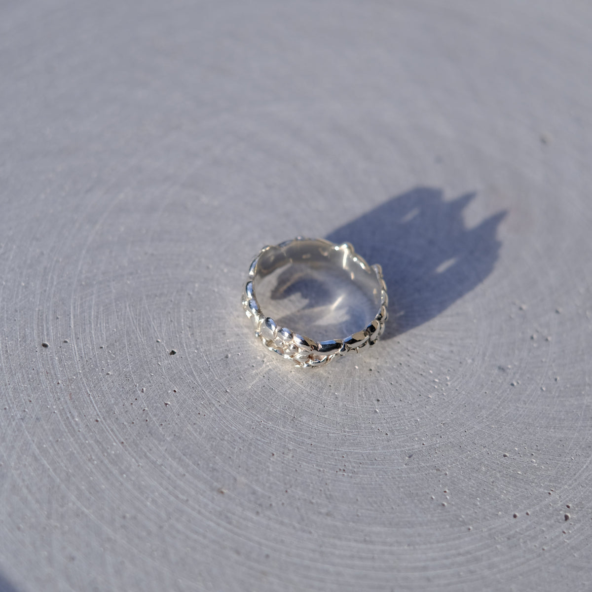 A clear faceted gemstone ring with a textured silver bezel, resting on a light gray textured surface with a sharp shadow.