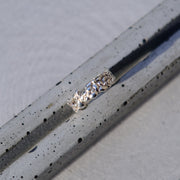 A silver ring with a textured, hammered band, resting diagonally on a speckled gray concrete surface.