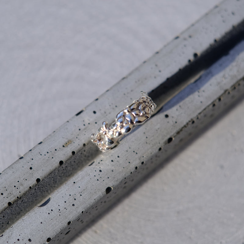 A silver ring with a textured, hammered band, resting diagonally on a speckled gray concrete surface.