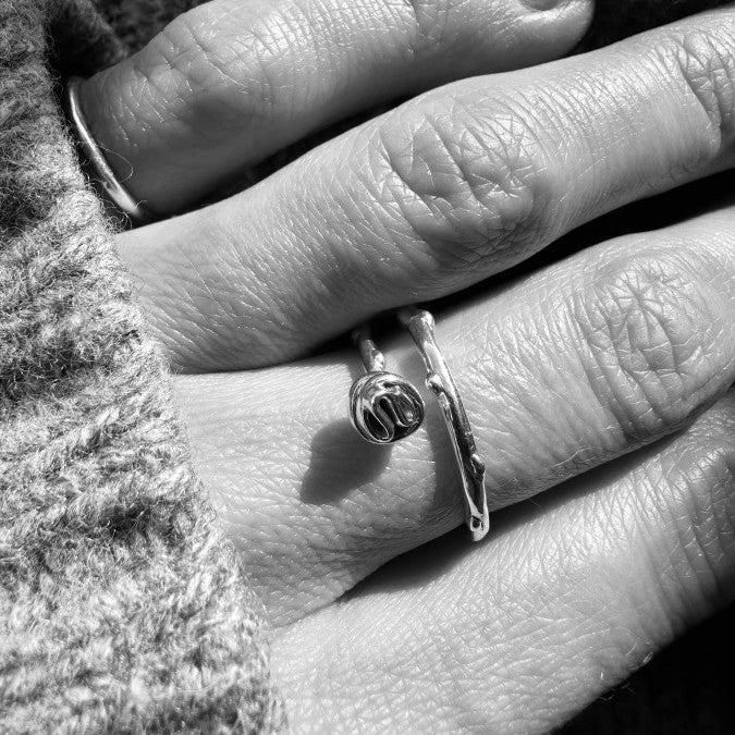 A close-up black and white photograph of a person's hand wearing silver ringwith a small round bead, set against a knitted gray sweater.