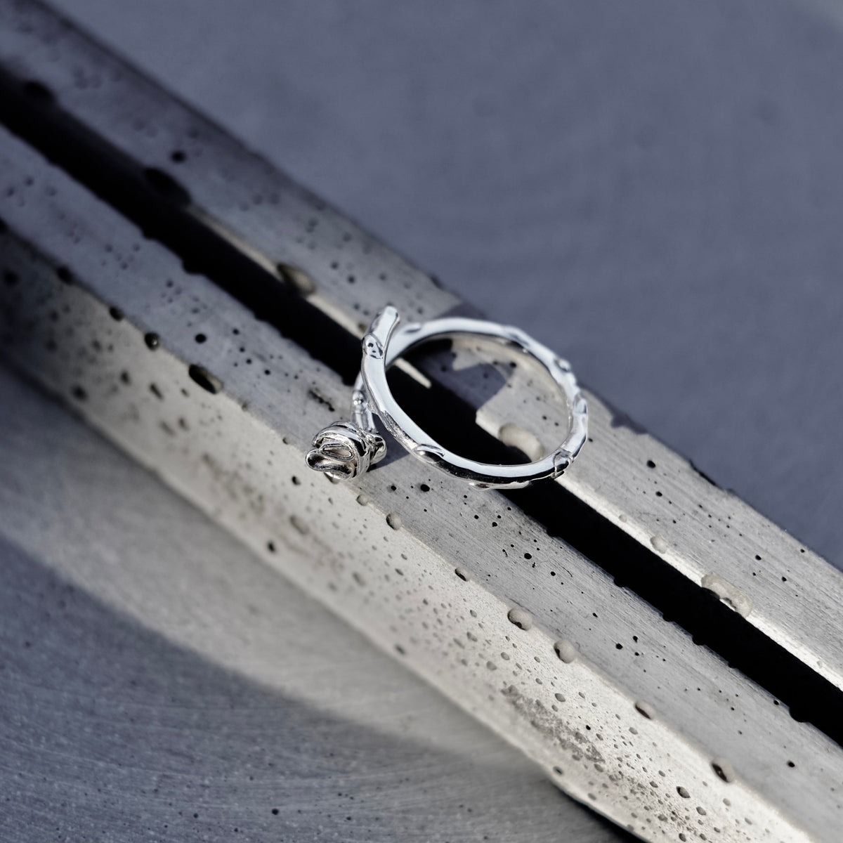 A silver textured ring with a small dangling bead detail, resting on a dark metallic surface.