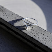 A silver-toned textured ring with a small clear stone, resting on a dark gray concrete ledge in bright sunlight.