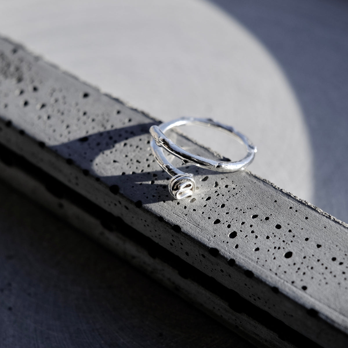 A silver-toned textured ring with a small clear stone, resting on a dark gray concrete ledge in bright sunlight.