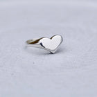 A polished silver heart-shaped ring with a smooth, reflective finish, resting on a textured light gray surface.