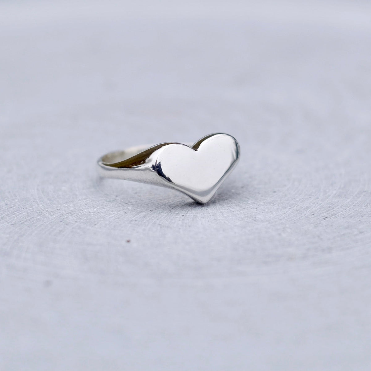 A polished silver heart-shaped ring with a smooth, reflective finish, resting on a textured light gray surface.