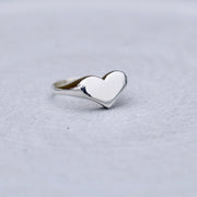 A polished silver heart-shaped ring with a smooth, reflective finish, resting on a textured light gray surface.