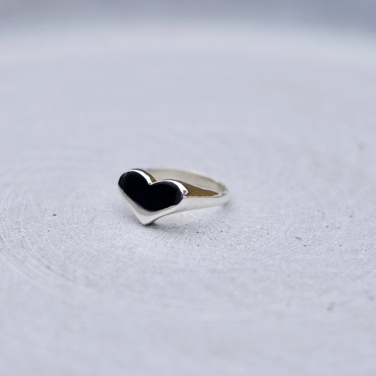 A silver heart-shaped ring, resting on a textured light gray surface.