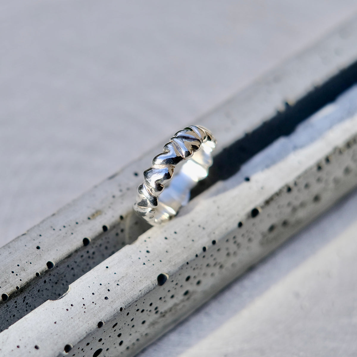 A silver ring with a textured, ridged band, resting on a speckled gray concrete surface.