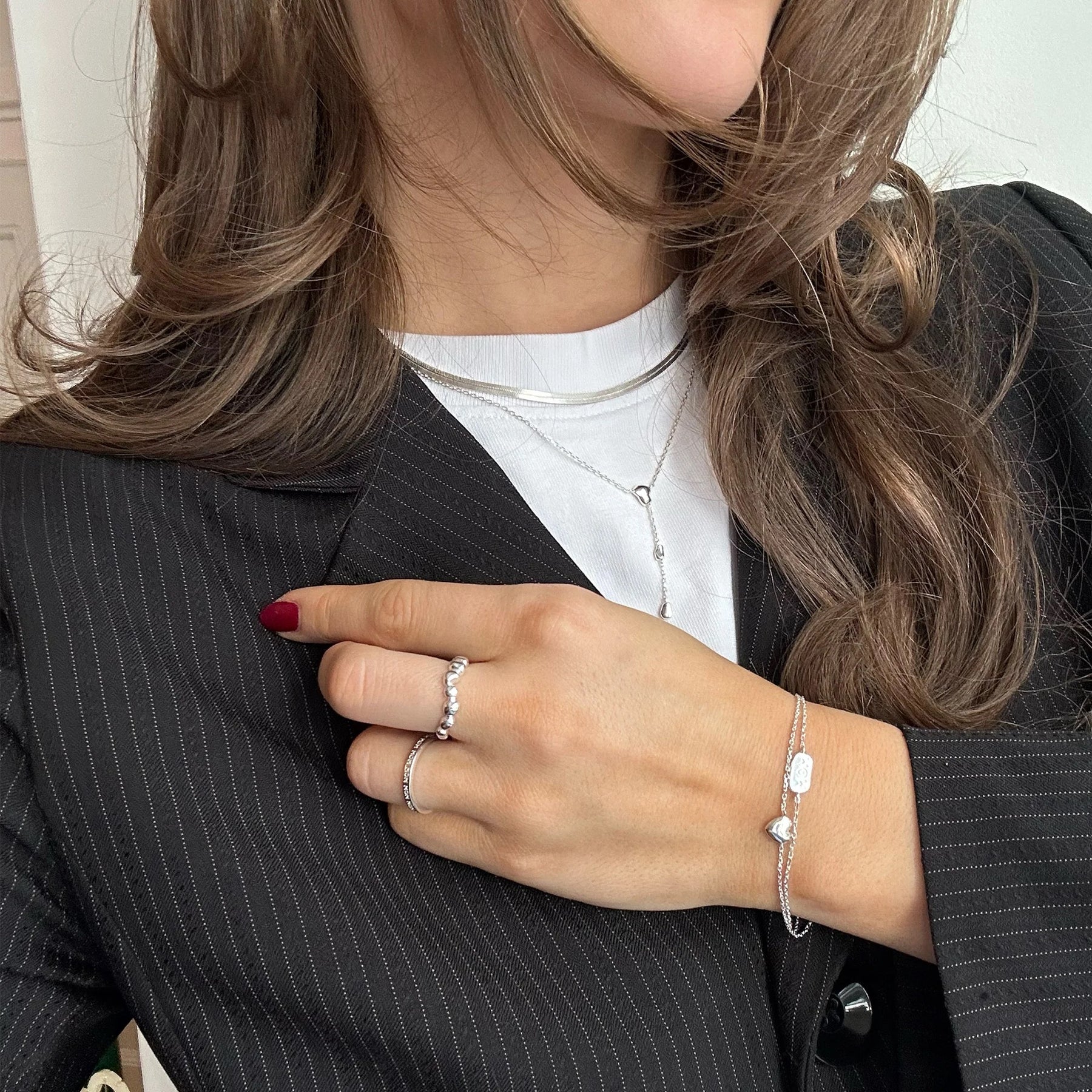 A close-up lifestyle shot of a woman wearing delicate silver jewelry, including a silver chain bracelet with white beads, silver rings, and a silver necklace, styled with a black pinstripe blazer and white t-shirt.