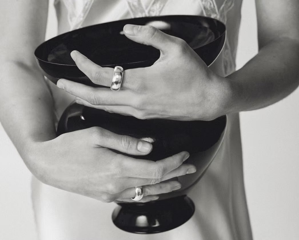 A close-up black and white photograph of a person holding a glossy black bowl with both hands, wearing silver rings on their fingers.