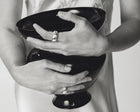 A close-up black and white photograph of a person holding a glossy black bowl with both hands, wearing silver rings on their fingers.
