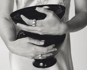 A close-up black and white photograph of a person holding a glossy black bowl with both hands, wearing silver rings on their fingers.