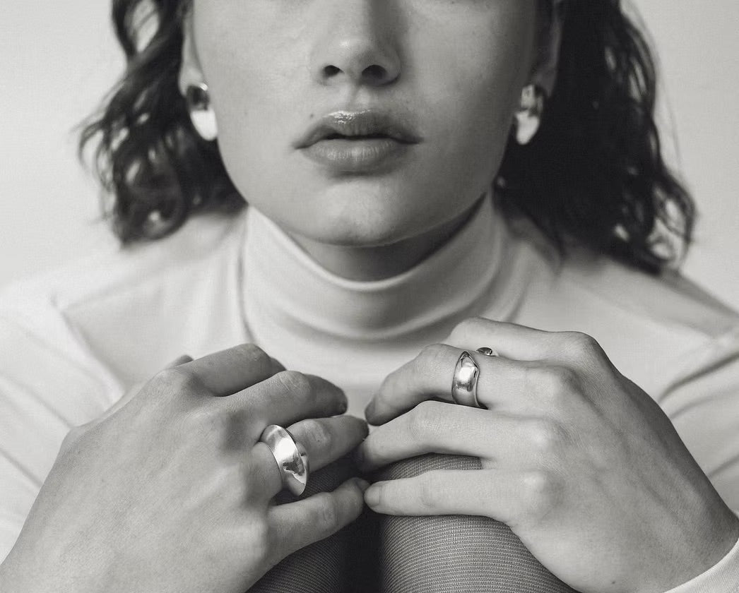 A close-up black and white photograph of a model wearing two wide, polished metal rings on their fingers, paired with a high-neck turtleneck top.