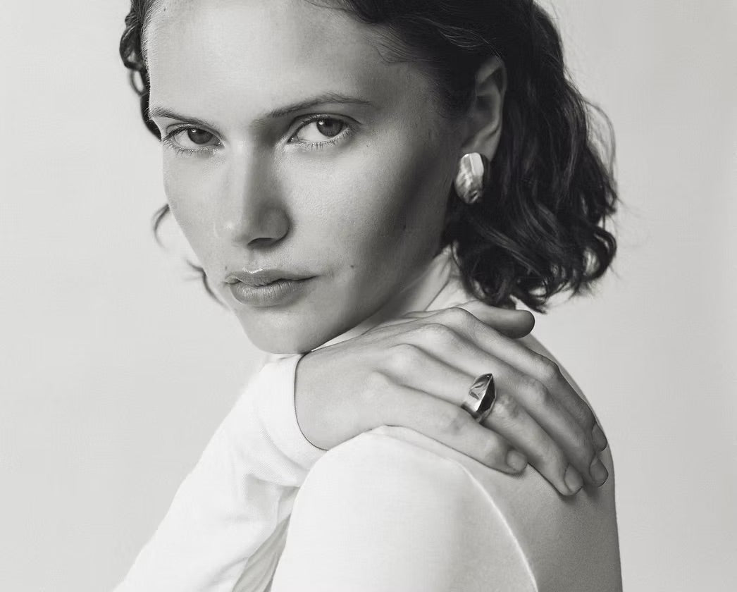 A black and white portrait of a woman with short dark hair, wearing a white top and jewelry including a ring and earring, with her hand resting on her shoulder.
