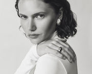 A black and white portrait of a woman with short dark hair, wearing a white top and jewelry including a ring and earring, with her hand resting on her shoulder.