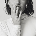 A black and white close-up portrait of a person wearing a wide silver band ring on their finger, with their hand resting against their lips.