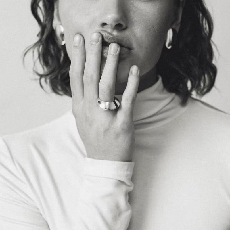 A black and white close-up portrait of a person wearing a wide silver band ring on their finger, with their hand resting against their lips.